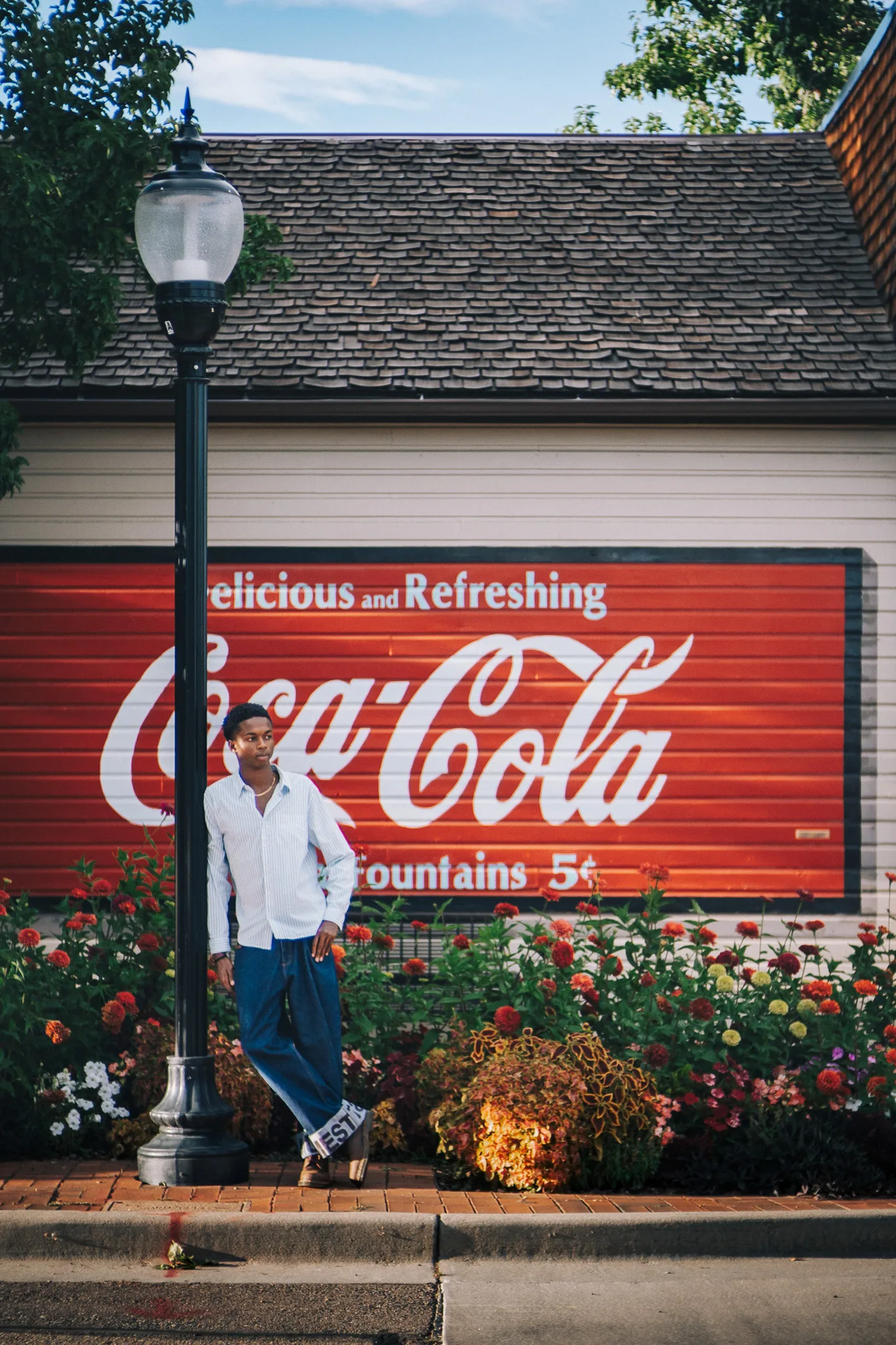 Charlie leans on a lamp post in front of a vintage Coca-Cola mural and a bed of flowers.