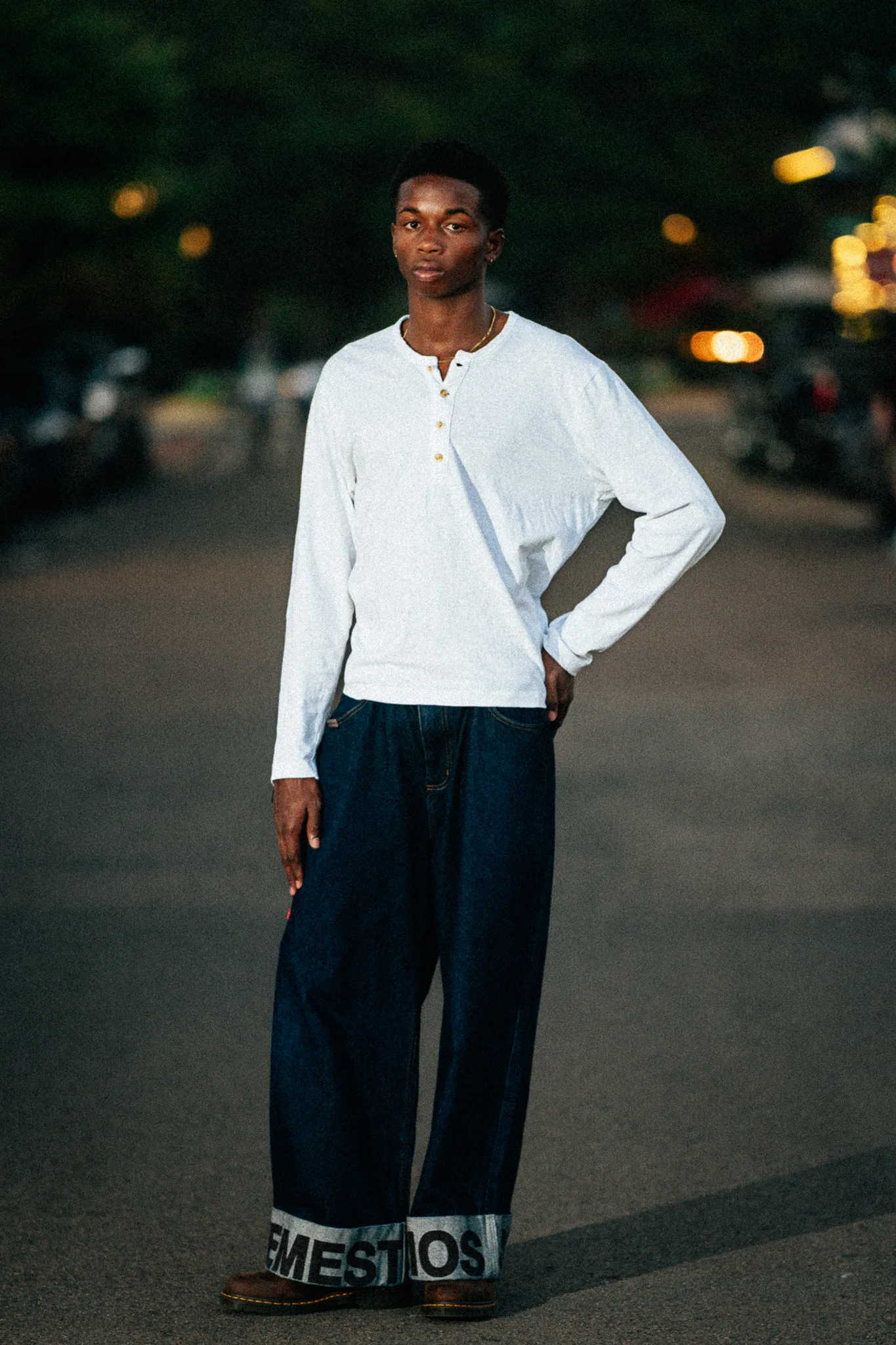Charlie stands in the street at dusk with one hand on his hip, lights glowing behind him.