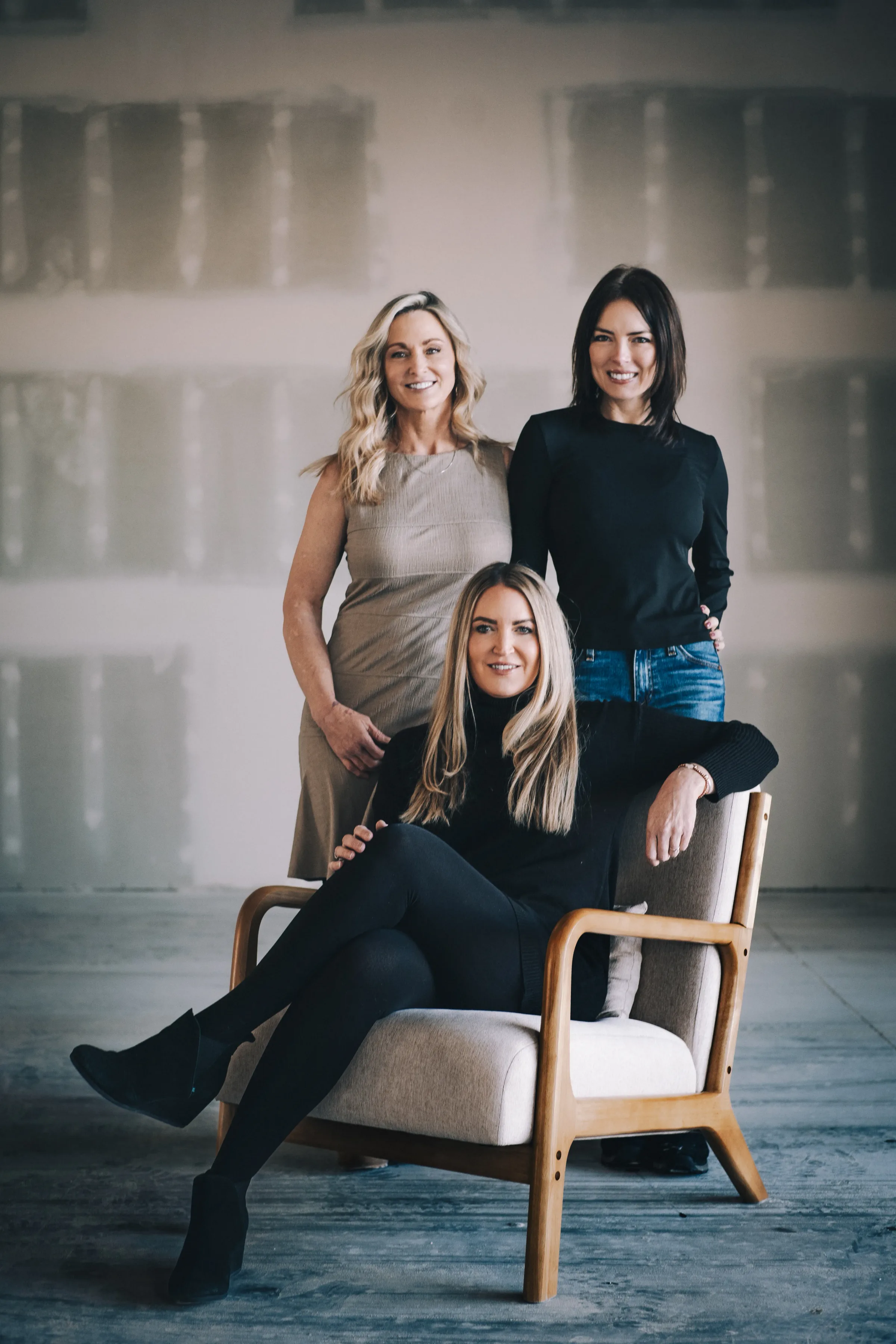 Three FACE Colorado team members pose around a wooden armchair in an open studio, two standing and one seated.