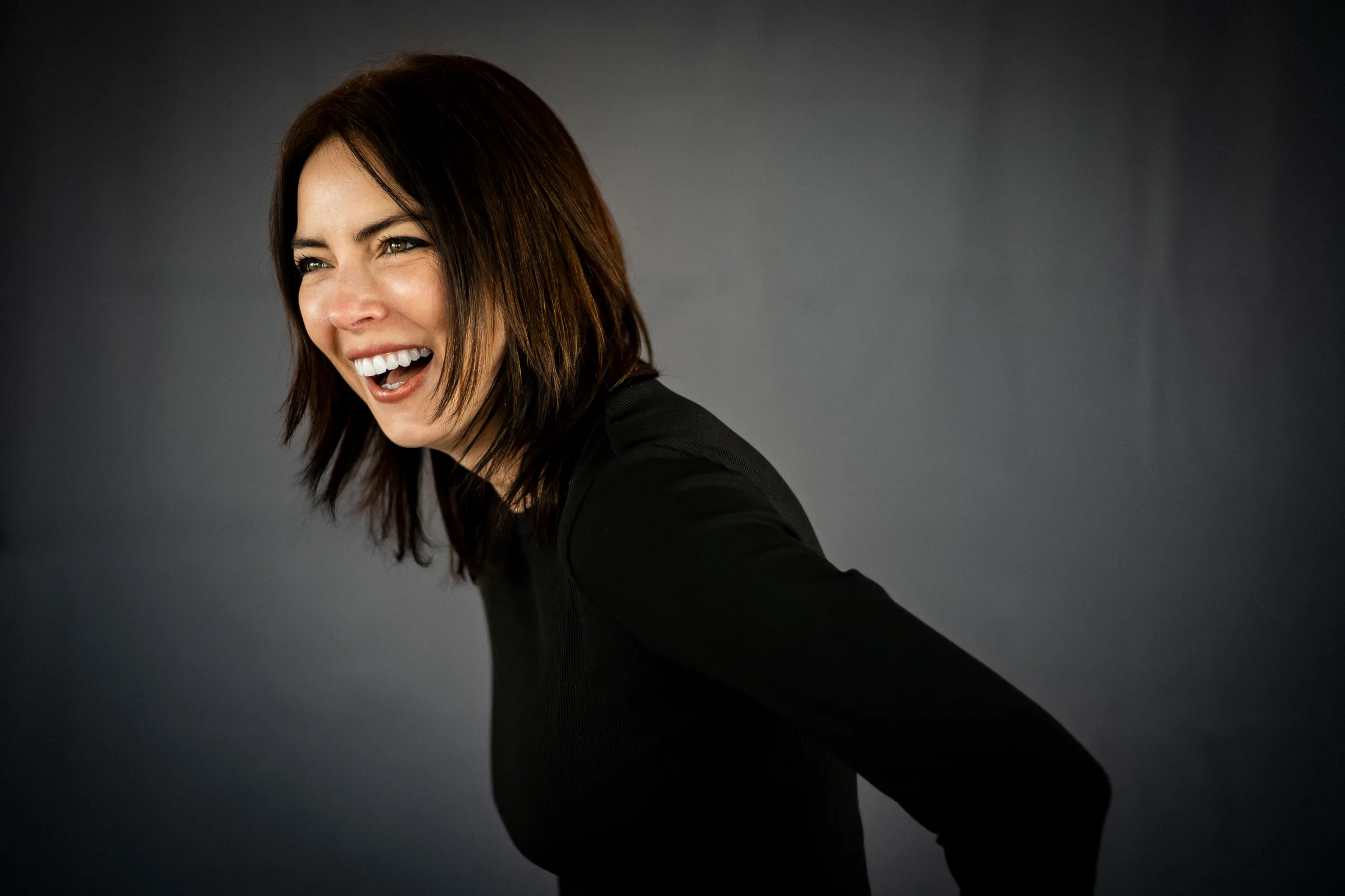 A FACE Colorado team member in a black long-sleeve top laughs while glancing to the side against a moody grey backdrop.