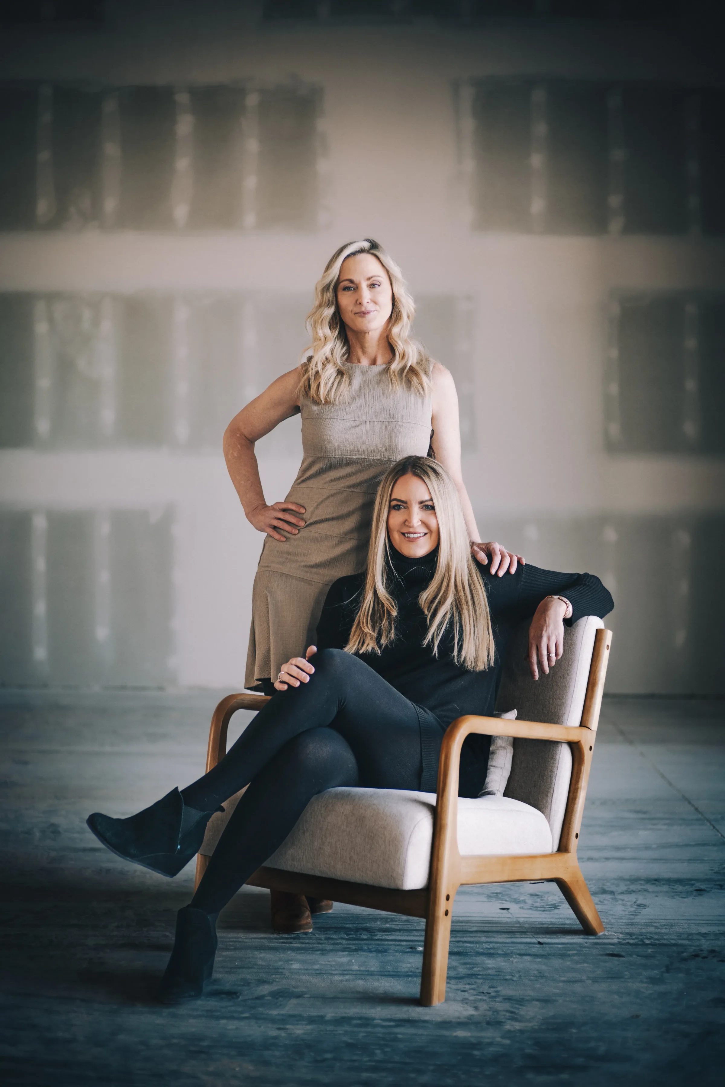 Two FACE Colorado team members pose with a wooden armchair in an open studio, one standing in a taupe dress and one seated in black.