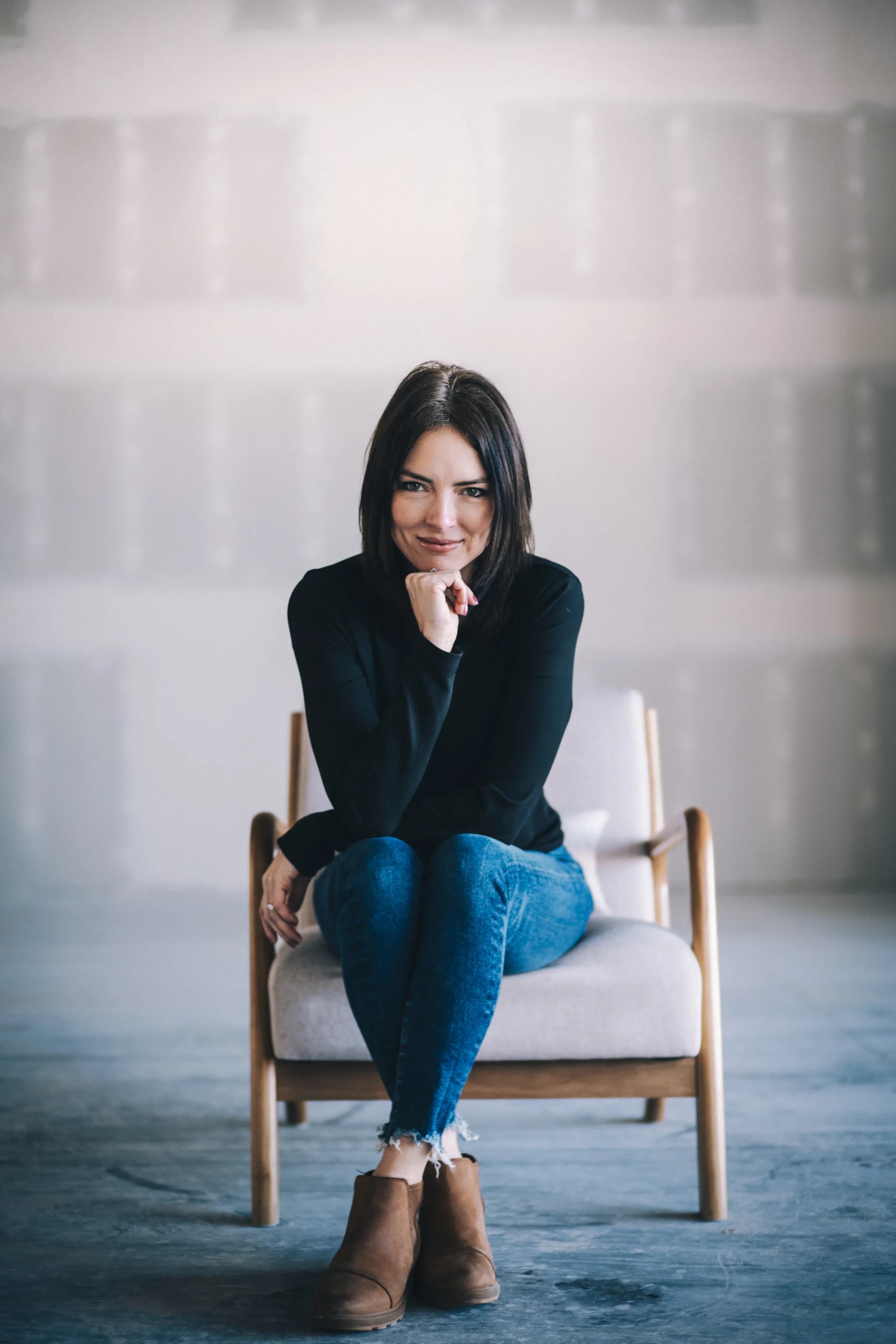 A FACE Colorado team member in a black top and jeans sits in a wooden armchair with chin resting on one hand.
