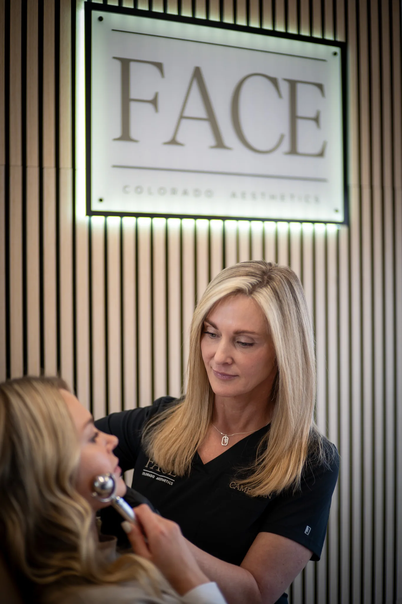 A FACE Colorado nurse in a black scrub top glides a metal facial globe along a client's jaw beneath the illuminated FACE sign.