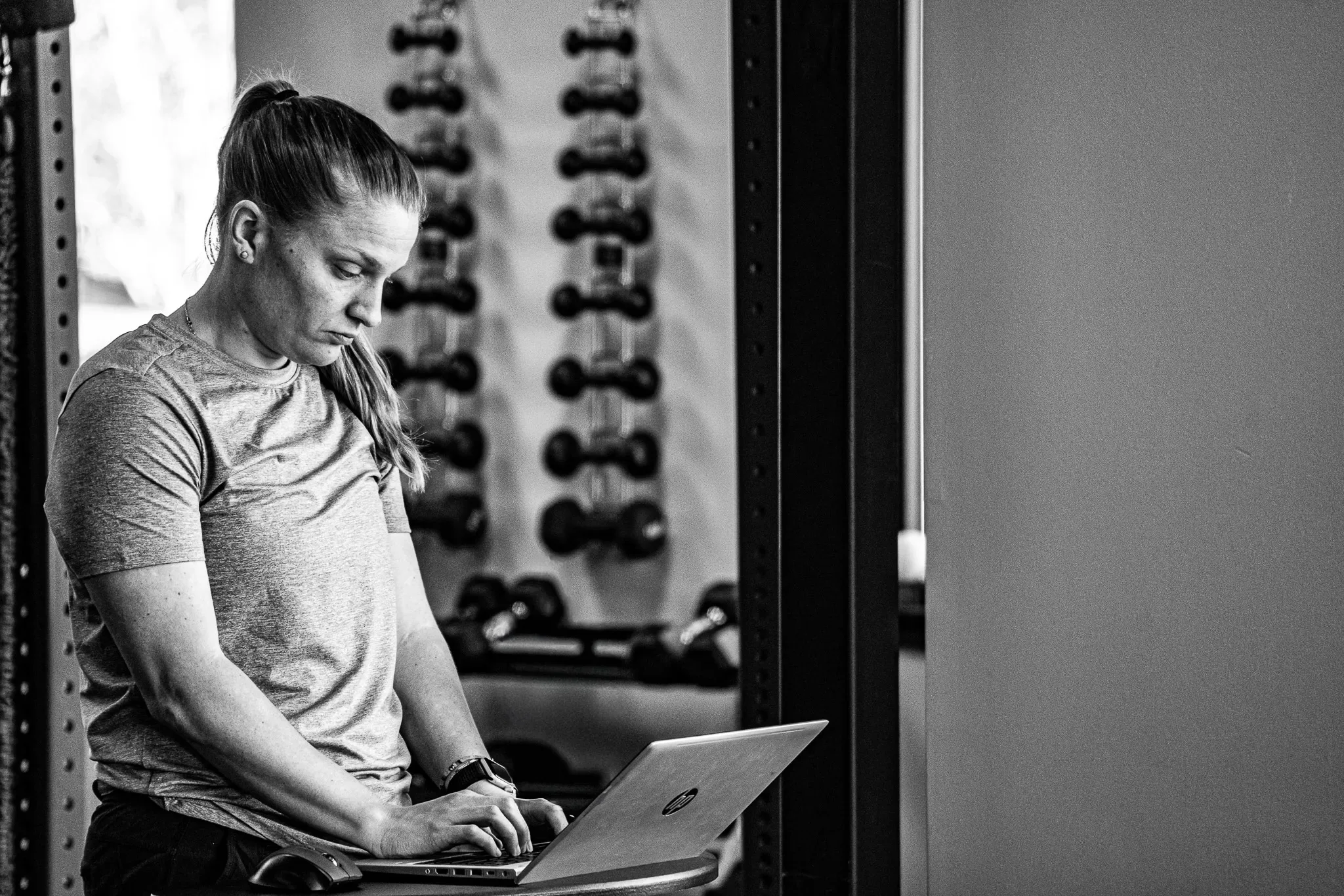 Black-and-white view of a person typing on a laptop beside a dumbbell rack in an IRG Hand & Physical Therapy clinic gym.