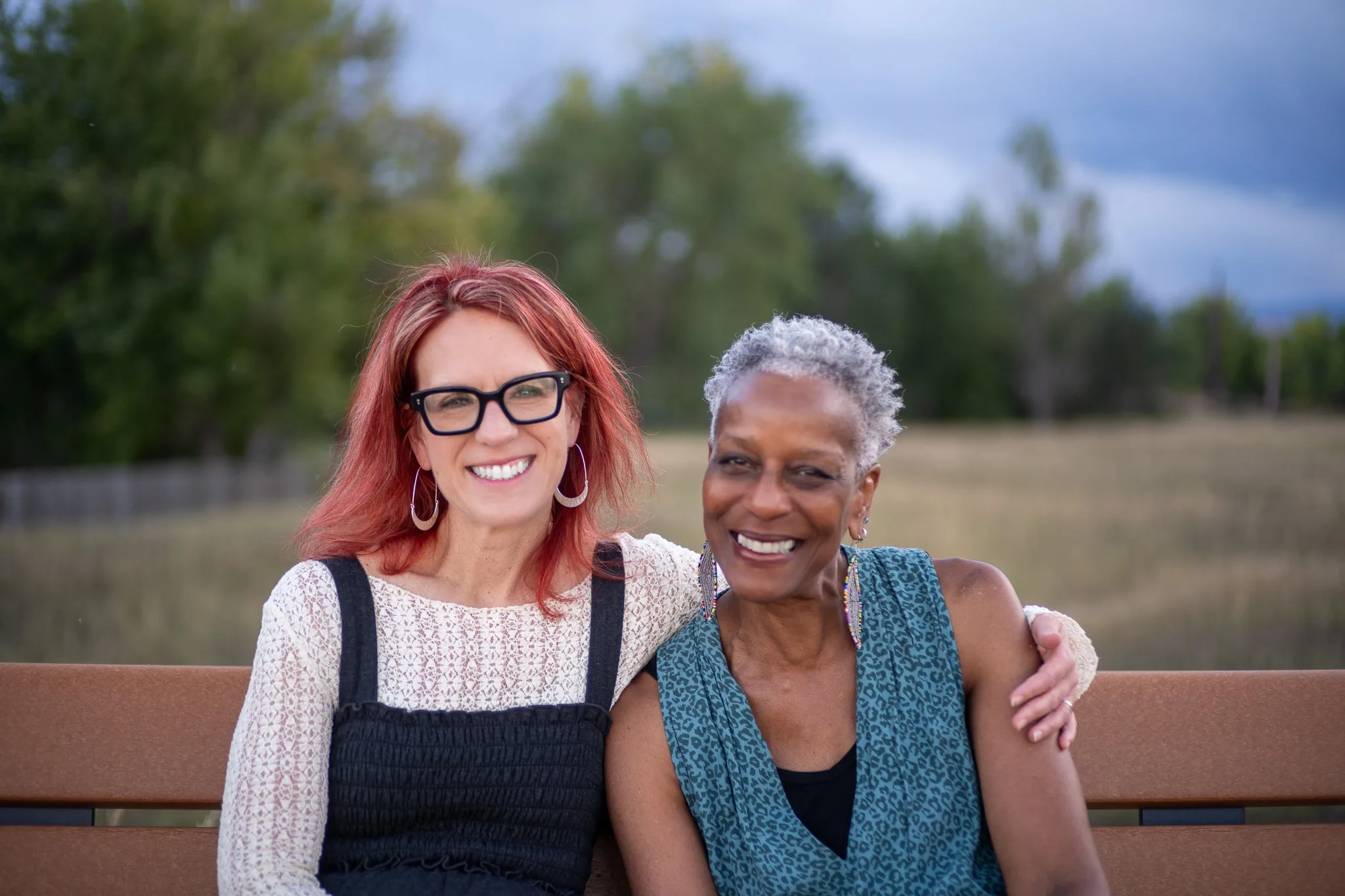 Amanda Cherry and Norma Johnson, co-founders of Who Tells the Story, smile arm in arm on a park bench in a field.