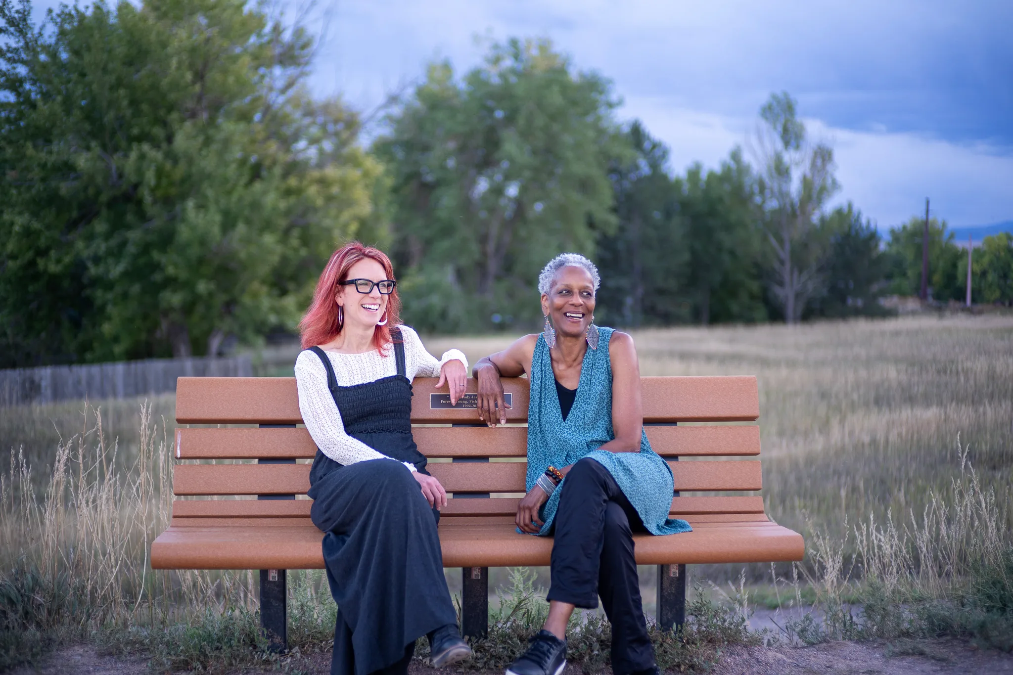 Who Tells the Story co-founders Amanda Cherry and Norma Johnson laugh together on a park bench in an open field.