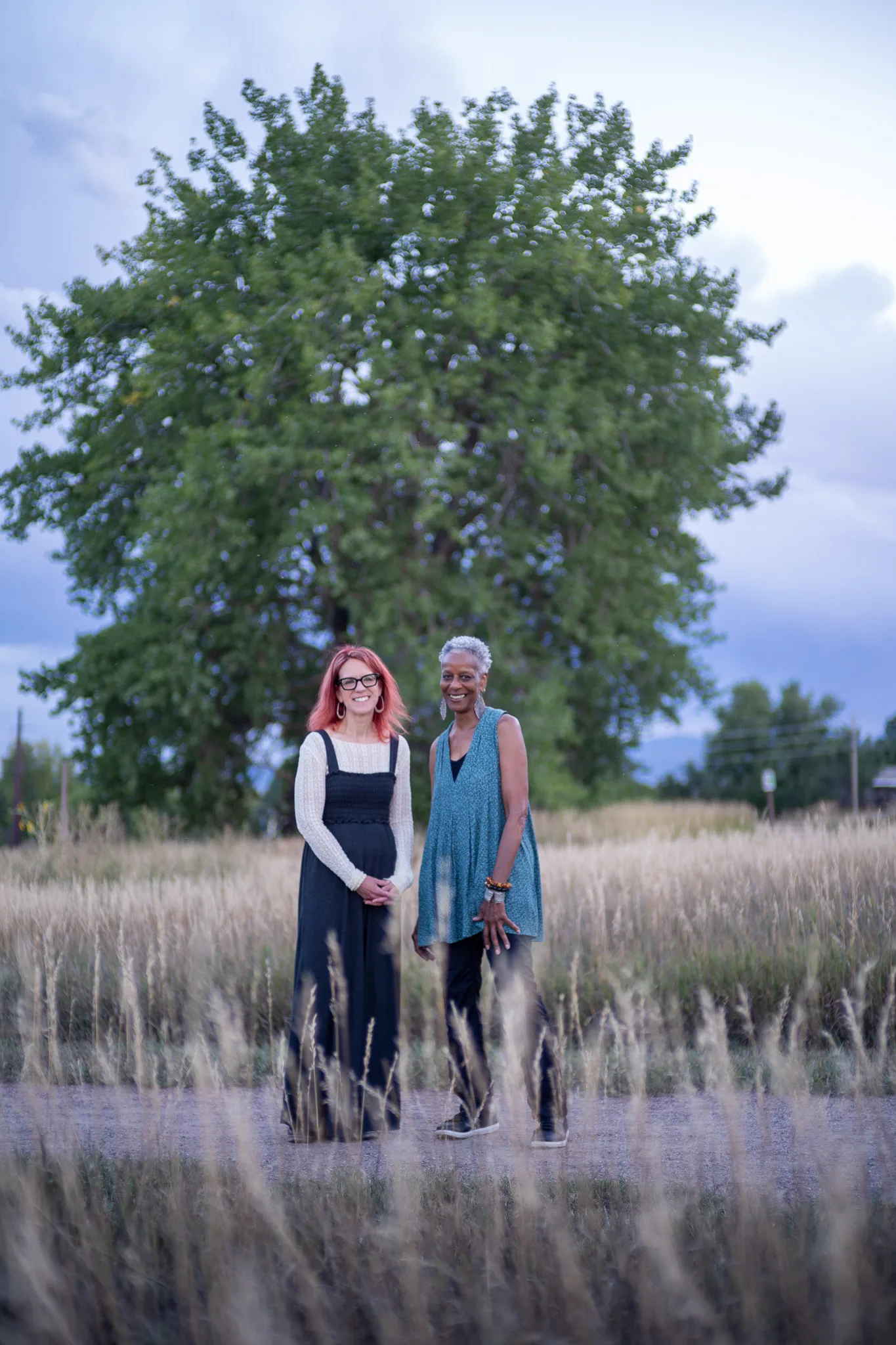 Amanda Cherry and Norma Johnson of Who Tells the Story stand smiling on a path through tall grass in front of a large tree.