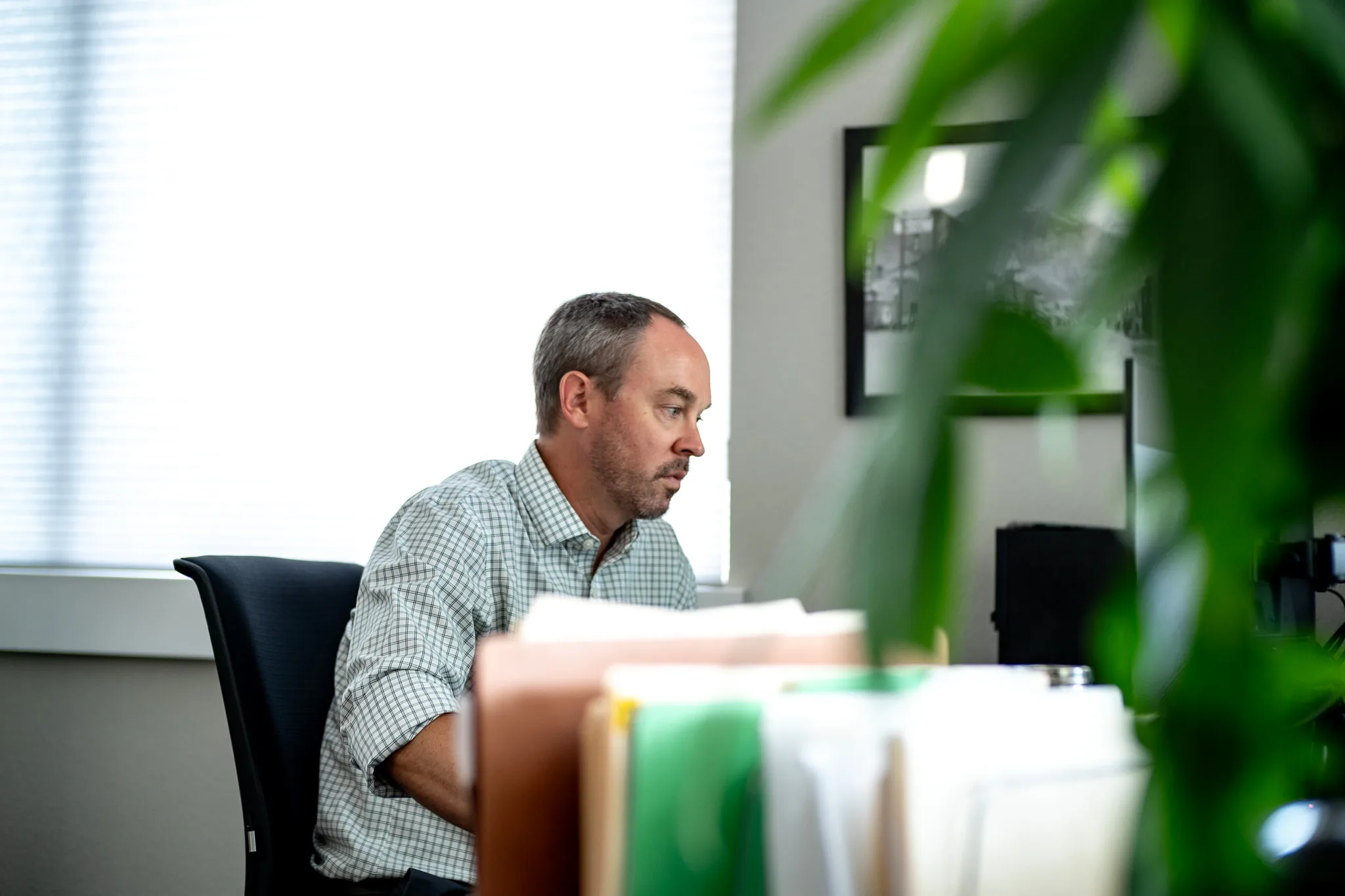 Seen past a leafy plant, a staff member in a checked shirt works at a desk stacked with file folders in a Wick & Trautwein office in Fort Collins.